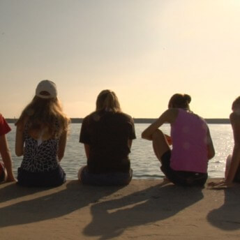 Still from Home Again. Five adolescent girls, seen from behind, are sitting on a water's edge. They are illuminated from the front by the sun, which casts long shadows behind them.