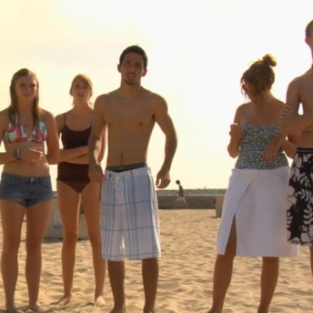 Still from Home Again. A group of six adolescents in beachwear are standing next to each other on sand. The majority are looking in the same direction offscreen.