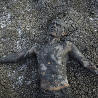 Still from Grit. A man is completely covered in a thick layer of grey mud. He is laying faceup on a ground covered in dried, cracked mud of the same color, and he almost blends in.