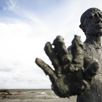 Still from Grit. A man covered in a thick layer of grey mud, is standing against a light blue sky, with his right hand outstretched toward the camera.