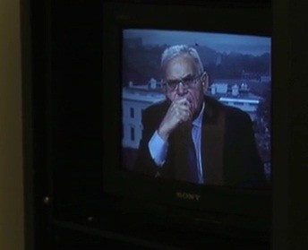 Still from My Favorite Neoconservative. A man on a television set is looking at the camera with his hand over his mouth, he is wearing glasses.