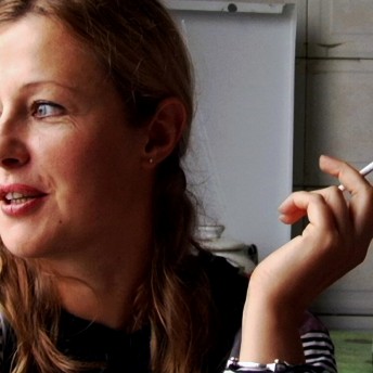 Still from My Perestroika. A woman is talking and looking off camera with a lit cigarette in her left fingers.