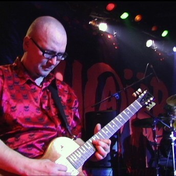 Still from My Perestroika. A man wearing glasses plays an electric guitar in front of a man playing drums on a drumset.