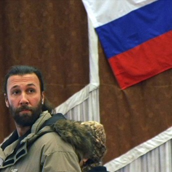 Still from My Perestroika. A man in a winter jacket stands in front of a wall. There is part of the Russian flag visible.