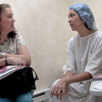 Two women talking to each other. One of them is in a hospital robe and wears a bouffant hat. The other one wears a t-shit and jeans, she has a bag in her lap and opened magazines on top of it.