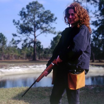 Julie Kahn stands slighty far away from the camera, she is standing on a patch of grass in front of a pond. She is turned to the side away from the camera with her head turned to the left to face the camera. She has slightly longer than shoulder length hair that is brown and curly. She is wearing jeans and a blue jacket with a yellow bag hanging at her left hip. She is holding a shotgun pointed down towards the ground.