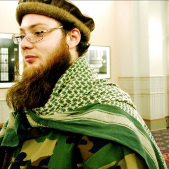 Still from (T)ERROR. A man with a beard faces to the right. He has a large beard and is wearing all green.