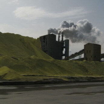 A still from The Factory and Me. A factory is nestled next to a series of green hills on the opposite side of a river. White smoke billows from the pylons.