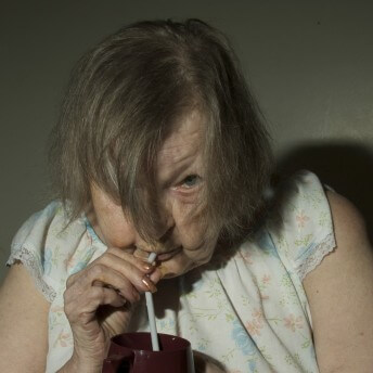 Still from The Patron Saints. A person stands in front of the camera wearing a sleeveless nightgown. They hold a straw to their mouth and peer up at the camera from between their hair which falls down over each side of their face.