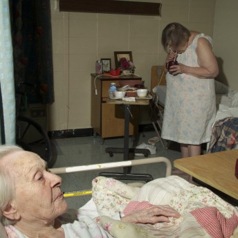 Still from The Patron Saints. Two people are in a sterile-looking room. The walls are made of large white brigs. There are two beds in the room, two bedside tables, and limited other furniture. One person stands in front of their bed in the background, wearing a night dress and sipping a drink through a straw. The other person in the foreground lays on their bed, underneath a quilt.