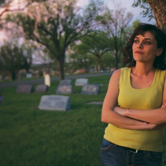 Still from Uranium Drive-In. A woman in a yellow tanktop stands in a cemetery, leaning against a tree.