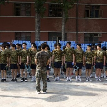 Still from Web Junkie. A lined-up group of boys wearing camo t-shirts all facing the same direction. Behind them is a windowed building. Color photograph.