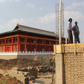 Two men are standing on the top of a construction building. In the background there are is a red Buddhist temple.