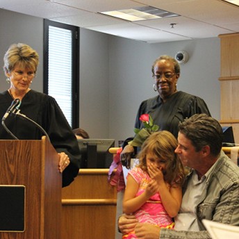Still from Tough Love. A man has a little girl sitting on his lap, she is covering her face with her hands. Next to them is a judge, and behind them, there is another judge.