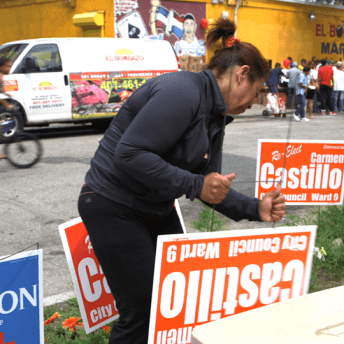 A woman placing posters on the grass that say "Re-elect Carmen Castillo"