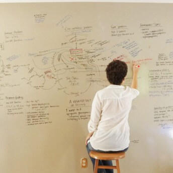 Still form Unrest. A woman sits on a stool facing a wall with a whiteboard full of writing.