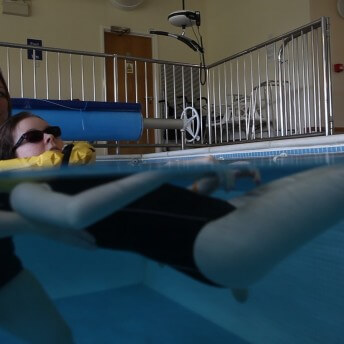 Still form Unrest. A woman in a pool is holding another young woman floating in a pool. The image is half underwater.