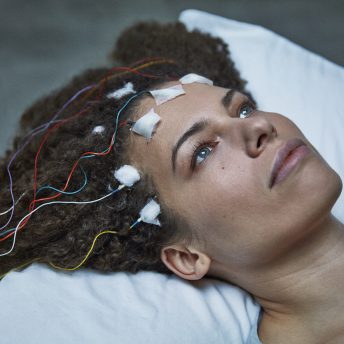 Still from Unrest. A woman's head is lying on a pillow with sensors taped to her forehead, the wires laying on her hair.