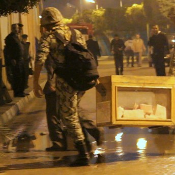 Still from The Vote. People in the street watch as soldiers carry boxes with votes inside of them.