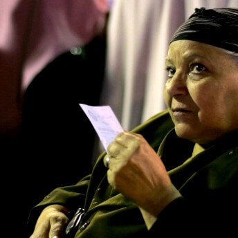 Still from The Vote. A woman wearing a scarf on her head holds a piece of paper in her hands. She looks up from the camera.