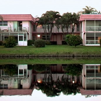 Still of the film Kings Point. A full shot of two pink houses next to each other, there are two trees between them. Their reflection is seen in the body of water in front of them.