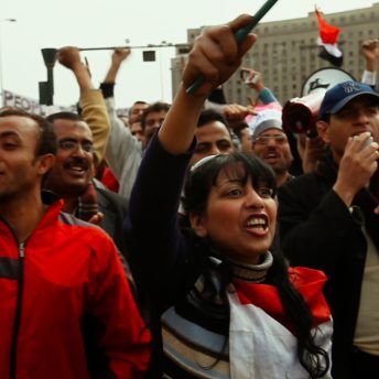 Still from The Trials of Spring. A large crowd of people with their hands raised. Color photograph.