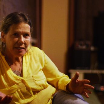 Still from The Trials of Spring. A person sitting down speaking with animated hands. Color photograph.