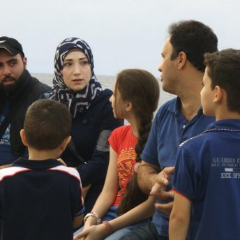 Still from It Will Be Chaos. A woman in hijab and a man, seated next to each other, are talking to another man. They are seated on a ledge with water visible behind them. There are four kids of varying ages around them.