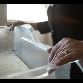 Still from No Mas Bebes (No More Babies). A person stands beside an empty hospital bassinet basket with their hands resting on the sides.