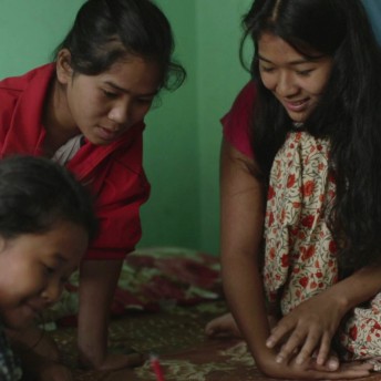Still from Even When I Fall. Three adolescent girls are sitting on the ground. They wear color, patterned clothing. Two of the girls are watching the third girl in the foreground as she is writing.