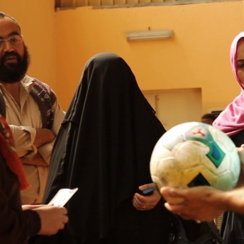 Still from Freedom Fields. A group of people inside a building whose walls are a bright orange color. The focus of the image is the woman in the middle who wears a black burqa and holds a phone in one hand. She is surrounded by four other women who are conversing with each other. A man in the foreground is wearing a blue-striped jersey and is holding a soccer ball.
