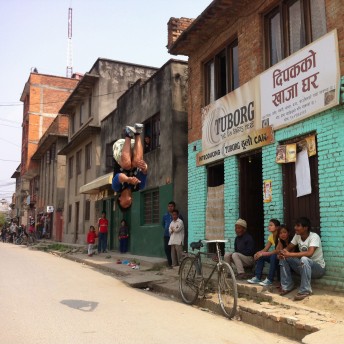 Still from Even When I Fall. A street scene. A row of buildings are on the right side of the street, with the bricks of building in the foreground painted a bright green-blue color. A man does a midair somersault in the middle of the street. Some of the adults and kids who are on street watch him. The sign on one of the buildings is written in Nepali.