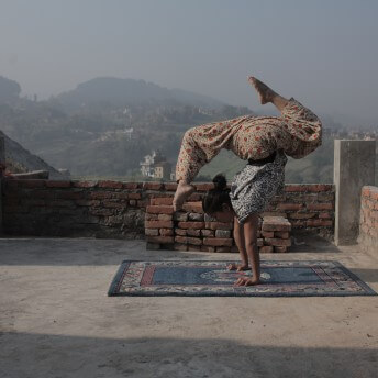 Still from Even When I Fall. A young woman is upside down on a blue Persian-style rug. She is balancing on her arms, with one leg lifted in front of her head, and the other leg in the air. She is wearing a black-and white patterned shirt and brightly-partterned pants. She is on a rooftop; houses, fields, and forested hills are seen in the distance behind her.