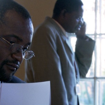 Two men in business attire. One of them holds some papers and the other one is talking via cellphone.