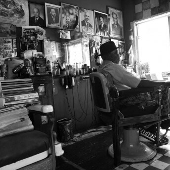 A still from The Barber of Birmingham. A man sits in a barbers chair, turned away from the camera. The wall behind him is covered in headshots, and various ephemera are stacked on the surfaces around him. Photo in black and white.