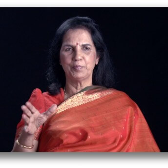 Suniti Solomon speaking to the camera in an orange Indian Sari.