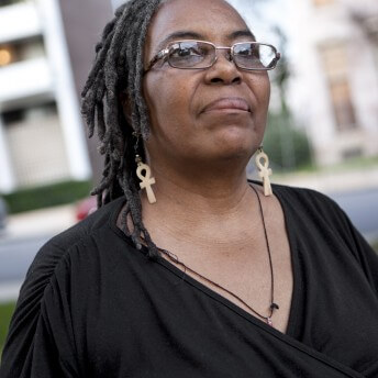 A still from The Graying of AIDS. A person wearing a black shirt and glasses facing the camera looks off into the distance. They wear a pair of wooden ankh earrings and a thin black necklace.