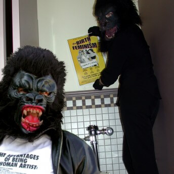 Two people in gorilla suits and face masks. One is sitting and the other stands behind them touching a poster on the wall.