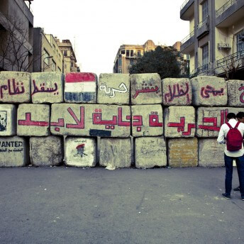 Large stacked blocks forming a wall covered in spray painted Arabic text. Two people are standing to the right. Residential buildings stand behind the wall.