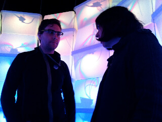 Still from The Mouse That Roared. Two people stand looking at each other. They are in a dark room with a wall of plastic containers stacked behind them. The plastic containers are lit up with bright, neon, technicolor, and white lights.