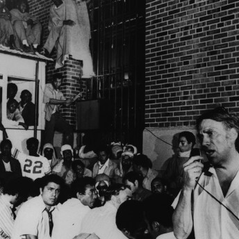 A group of men sit and stand close to one another beside a brick building. One man is speaking into a public announcement system. Black and white photograph.