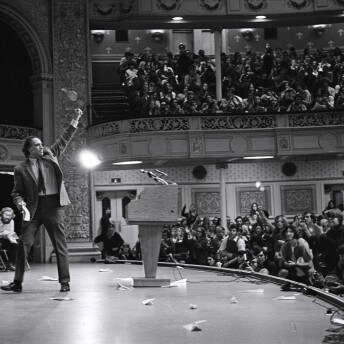 William Kunstler raises a fist to the sky on a stage in front of a podium. The crowd of people in the background also stands. Black and white photograph.