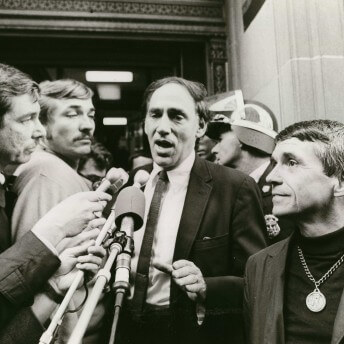 William Kunstler speaking into many microphones, surrounded by several people. Black and white photograph.