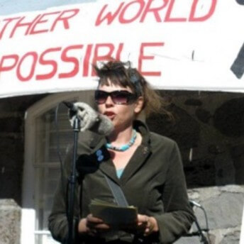 Still from The Mouse That Roared. A person wearing sunglasses stands outside in front of a microphone. They are holding a small stack of papers in their hands. Behind them, a large banner reads: "-nother World Possible".