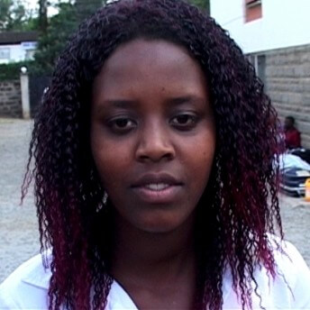 Still from Pushing The Elephant. A woman stands in an exterior in front of the camera, she is not looking at the camera. There are people and suitcases in the background.