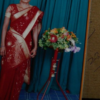 A framed portrait of a woman in a red sari standing beside a bouquet of flowers placed on a cardboard surface with handwriting that says "23 yr" to the right of the frame.