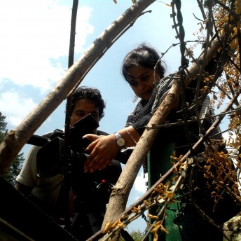 Production still from I Never Left. A woman positions a camera on a tripod. She is seen from a low angle in between branches. A man is looking on.