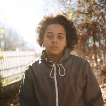 Still from Tre Maison Dasan. A young teenager looks at the camera. They are in a exterior and the sun shines through the leaves of the trees behind them.