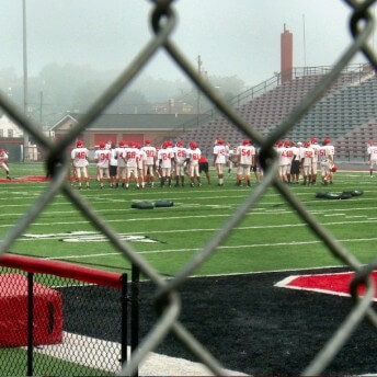 Still from Roll Red Roll. The shot was taken through a steel fence in the foreground, in the background a football team with white jerseys and red helmets practices on a football field.