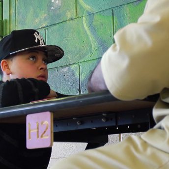 Still from Tre Maison Dasan. A young boy wearing a cap is sitting on a bench, in front of a person wearing a prison uniform.